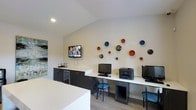 This single-floor business center offers a well-appointed workspace designed to meet both professional and recreational needs. The room features a sleek, continuous white countertop workstation accommodating multiple computer terminals and a shared printer, making it a practical hub for work and connectivity. Dark cabinetry lines the adjacent wall, providing ample storage while housing a small beverage station with a mini-fridge, adding a convenient amenity for users. A wall-mounted flat-screen television is positioned above the cabinetry, offering an easy source of information or entertainment. The vaulted ceiling enhances the sense of openness in the space, while recessed lighting provides consistent, functional illumination throughout. The light hardwood flooring contributes to a clean, contemporary aesthetic. A standout design element is the collection of decorative circular wall pieces in varying sizes, finished in rich tones of blue, red, orange, and black, which add an artistic focal point to the otherwise neutral palette. A framed abstract artwork on the adjacent wall further enriches the room's visual character. Overall, the space balances functionality with thoughtful design details.