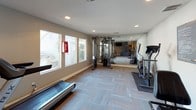 This fitness center is a thoughtfully designed single-level workout space that accommodates a variety of exercise needs. The room features neutral gray walls, recessed lighting throughout, and a patterned carpet tile floor that adds visual interest while providing a functional surface for workouts.

The main exercise area is generously proportioned, housing a treadmill, elliptical, stationary bike, rowing-style machine, and a multi-function cable resistance station. A large mirrored wall on one side visually expands the space and allows users to monitor their form. A row of windows along one wall allows natural light to fill the room and provides outdoor views, contributing to an open and energizing atmosphere.

Additional amenities include a wall-mounted flat-screen television, a wall-mounted fan for ventilation, and a wall-mounted sanitizer dispenser. A stability ball and seating chair are also present. A dedicated door leads to an adjacent secondary room, which offers additional equipment storage or workout space.

The overall ambiance is clean, functional, and practical, making this fitness center a capable and well-appointed facility for a broad range of exercise routines.