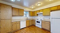 This single-floor residence offers a straightforward and functional layout designed for comfortable everyday living. The kitchen is well-equipped, featuring warm oak-toned cabinetry that extends to the ceiling for ample storage, light-colored countertops, and a suite of white appliances including a dishwasher, electric range, and refrigerator. A window with vertical blinds allows natural light to brighten the space, while hardwood-style flooring adds a clean, durable finish underfoot. A pass-through opening connects the kitchen to the adjacent living area, creating an open and connected feel between the two spaces. The living area is generously sized, finished with wall-to-wall carpet in a warm neutral tone, and features a ceiling fan overhead. Multiple bedrooms continue the carpeted theme, each with vertical-blind windows that let in natural light. One bedroom includes a sliding-door closet for convenient storage, while another features its own ceiling fan. The bathroom is neatly appointed with a single-sink vanity topped with a wide mirror, hardwood-style flooring, and a full tub-shower combination with tiled surround. Throughout the residence, the neutral color palette and clean finishes provide a versatile backdrop well-suited to a variety of interior styles.