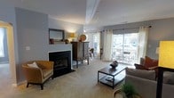 This thoughtfully designed single-floor space offers a welcoming and functional layout centered around a stylish living area. The room is anchored by a sleek fireplace with a black surround and a decorative mantel, adding warmth and a focal point to the space. Soft, neutral-toned carpet runs throughout, complementing the light gray walls and creating a calm, cohesive atmosphere. Natural light pours in generously through large sliding glass doors dressed with floor-length white curtains, which open onto an outdoor patio furnished with seating — extending the living space beyond the interior. The living area is well-furnished with a mix of modern and rustic elements, including a metal-framed coffee table with a lower shelf for storage, a caramel leather accent chair, and a vintage-style wooden cabinet that adds character and depth. A framed mirror above the mantel reflects the light, enhancing the sense of openness. Recessed lighting and track fixtures provide supplemental illumination, while decorative accessories — including ceramic vessels and woven accents — contribute to the curated, lived-in feel. Overall, the space balances practicality with aesthetic appeal, offering a bright, inviting environment with indoor-outdoor connectivity.