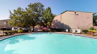 This single-story property showcases a well-maintained outdoor swimming pool as its centerpiece amenity. The generously sized pool features clear, turquoise water and is framed by a classic black iron fence that provides both security and a clean aesthetic boundary. A wide pool deck surrounds the water, offering ample space for lounge chairs and outdoor furniture, making it ideal for relaxation and recreation.

Mature, leafy trees line the perimeter of the property, providing natural shade and a lush, green backdrop that enhances the outdoor atmosphere. The open layout allows for abundant natural sunlight throughout the day, creating a bright and inviting environment. The building adjacent to the pool area features a combination of brick and neutral-toned siding, characteristic of durable, low-maintenance construction.

The overall ambiance of the outdoor space is open, airy, and communal, well-suited for warm-weather enjoyment. The combination of mature landscaping, a spacious pool, and thoughtfully arranged seating areas creates a welcoming retreat. The property's single-floor design contributes to an accessible and cohesive layout, ensuring the outdoor amenity is conveniently reachable from all areas of the building.