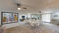 Located at 600 Valley Rd in Warrington, PA, this single-floor community space offers a well-appointed combination of a kitchen and living room designed for gathering and socializing. The open layout is unified by a striking wave-patterned carpet that flows throughout, lending the space a modern, welcoming aesthetic.

The main area features multiple round dining tables with sleek white chairs, providing ample seating for group events or casual gatherings. Ceiling fans are positioned throughout to ensure comfortable air circulation. A standout architectural detail is the full-wall mirrored panel along one side, which visually expands the room and amplifies the natural light filtering in through the windows.

The living room section is furnished with a comfortable leather sofa, a coffee table, and a wall-mounted flat-screen television, creating a relaxed lounge environment. Bold pop-art wall decor adds a vibrant, expressive character to the space.

The kitchen is equipped with wooden cabinetry, granite countertops, tile flooring, stainless steel appliances including a range and microwave, and a full-size refrigerator. A pass-through counter connects the kitchen to the main gathering area, making it ideal for serving during events. A workstation area with computers is also present, adding a functional utility component to this thoughtfully designed community space.