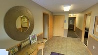 This single-floor space presents a functional and professionally designed interior well-suited for office or service-oriented use. The entryway sets a welcoming tone, featuring light tile flooring, neutral-toned walls with warm wood-trimmed accents, and a decorative circular mirror mounted above a small glass-topped accent table. Framed artwork adds a touch of personality to the reception area.

Light maple-finished doors line the entry corridor, providing access to adjacent rooms, while ADA-compliant accessibility signage indicates that the space is designed with inclusivity in mind. Flush-mounted ceiling lighting provides consistent, even illumination throughout.

Beyond the entry, a carpeted hallway extends further into the space, offering additional rooms and areas. The transition from tile to carpet creates a natural delineation between public-facing and more private or functional zones. The hallway is lined with framed artwork and benefits from overhead lighting that maintains a bright, navigable atmosphere.

Overall, the space conveys a clean, organized, and practical environment. The combination of neutral color palettes, warm wood accents, and accessible design elements contributes to an atmosphere that is both professional and approachable — well-suited for welcoming visitors and supporting daily operations.