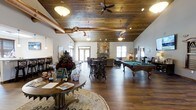 The Copper Creek Clubhouse is a thoughtfully designed single-floor gathering space that blends rustic charm with modern comfort. The interior is anchored by striking wood-planked ceilings with exposed copper piping and ceiling fans, lending the space a warm, lodge-inspired aesthetic. Rich hardwood flooring runs throughout, complementing the natural tones of the décor.

The open-concept layout accommodates a variety of activities and social settings. A welcoming reception area greets visitors with a curved wood desk, floral arrangements, and a chalkboard sign. High-top bar seating and casual lounge areas are arranged throughout the main space, providing flexible options for both small gatherings and larger events.

A full-sized billiards table serves as a centerpiece for recreation, while a cozy lounge area with comfortable seating surrounds a stone fireplace, creating an inviting focal point for relaxation. Natural light filters in through multiple windows and glass doors, brightening the interior and offering views to the outdoors.

Additional amenities visible throughout the space include gaming and activity stations along the walls. The overall ambiance is warm, social, and community-oriented, making this clubhouse a versatile hub for residents to connect and unwind.
