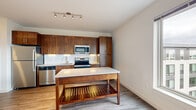 This single-floor unit presents a clean, modern aesthetic with a thoughtfully designed layout connecting living, dining, and kitchen areas. The kitchen is a standout feature, showcasing rich walnut-toned cabinetry extending to the ceiling, a white subway tile backsplash, and light granite countertops. A full suite of stainless steel appliances — including a refrigerator, range, microwave, and dishwasher — provides functional appeal. A freestanding wood island with an open lower shelf adds additional prep and storage space. Track lighting illuminates the kitchen area effectively. The open layout transitions seamlessly into the living space, where durable wood-look plank flooring runs throughout the main areas. Large windows in one room frame expansive views of a landscaped green space and adjacent buildings, filling the space with natural light. Bedrooms feature plush carpeting and include sliding-door closets with built-in shelving. The bathroom is finished with floor-to-ceiling white subway tile surrounding a full soaking tub and shower, complemented by large-format floor tiles, a wood-toned vanity, and chrome fixtures. Crisp white walls and clean lines throughout contribute to a bright, contemporary ambiance.