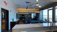 Located at 145 Westerly Rd in Ossining, New York, this single-floor amenity space is thoughtfully designed to offer an upscale, multifunctional environment with sweeping waterfront views. The open-plan layout seamlessly connects several distinct activity zones, creating a cohesive and inviting atmosphere throughout.

A striking sculptural chandelier serves as a dramatic focal point overhead, complementing a recessed tray ceiling with warm ambient lighting that sets an elegant tone. The space features a full-size billiards table positioned centrally, alongside a grand piano, making it well-suited for both recreation and entertainment. A dedicated lounge area furnished with contemporary chairs and tables provides comfortable seating, while a wall-mounted flat-screen television anchors a secondary gathering zone toward the rear.

Floor-to-ceiling windows and glass doors along the exterior wall flood the interior with natural light and offer panoramic views of the water beyond, enhancing the sense of openness. Neutral wall treatments, hardwood-style flooring, and sleek cabinetry contribute to the modern aesthetic. A white countertop surface in the foreground suggests a serving or bar area, adding further functional versatility to this well-appointed communal space.