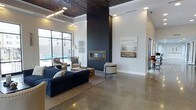 This contemporary single-floor space makes a strong architectural statement with its striking combination of materials and thoughtful design. The centerpiece of the room is a bold, floor-to-ceiling dark accent wall housing a sleek, framed linear fireplace, creating a dramatic focal point that anchors the entire space. Soaring ceilings clad in rich, dark wood paneling add warmth and texture, contrasting beautifully with the crisp white walls throughout. Polished concrete floors reflect light and lend an industrial-chic aesthetic that ties the modern design together seamlessly. Large, industrial-style windows along one wall flood the interior with natural light while offering views to an outdoor area. The lounge is furnished with a deep navy tufted sofa, neutral accent chairs, and layered area rugs, creating a comfortable yet sophisticated seating arrangement. An elegant chandelier with cascading metallic elements adds a refined decorative touch overhead. Curated wall art complements the neutral and navy color palette. A secondary area is visible in the background, suggesting additional functional space. Overall, the design balances industrial elements with polished, upscale finishes to create an inviting and visually dynamic environment.