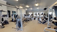 This fitness center is a comprehensive, single-floor facility designed to support a full range of workout routines. The space is generously sized, featuring vaulted and flat ceiling sections painted in a cool gray-blue tone, accented by white crown molding that adds a polished, refined feel throughout.

The main workout floor is divided into distinct functional zones. A dedicated cardio area houses a robust selection of treadmills, elliptical trainers, stationary bikes, and stair climbers, many equipped with individual monitors for entertainment. Adjacent to this, a strength-training section offers an extensive array of selectorized cable and weight machines targeting various muscle groups, with wall-mounted mirrors amplifying the sense of space and allowing users to monitor their form.

A separate free weights area features multiple racks of dumbbells ranging across various sizes, flat and adjustable benches, and a wall-mounted rack stocked with stability balls in multiple sizes. Elegant arched windows in this section flood the area with natural light, creating an inviting atmosphere.

Practical amenities include a water fountain station, ceiling fans throughout for air circulation, and overhead fluorescent lighting to ensure a well-lit environment. French door entries and large windows provide connectivity and natural brightness across the facility, making this fitness center a functional and welcoming space for all fitness levels.