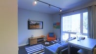 This single-floor space centers around a well-appointed living area designed with a modern urban aesthetic. The room features a sleek track lighting system mounted to the ceiling, providing adjustable, directional illumination across the space. A large wall-mounted flat-screen television serves as a focal point, flanked by light gray walls that lend a clean, contemporary feel. Colorful artwork adds visual interest above the seating area.

The living space is furnished with a bold orange armchair accented with a blue throw pillow, complemented by a blue sofa, creating a lively, contrasting color palette. A low-profile wooden media console sits beneath the television, offering practical storage. A glass-top coffee table anchors the seating arrangement atop a striking blue-and-white striped area rug, adding a nautical touch to the overall design.

A large floor-to-ceiling window along one wall floods the room with natural light and offers open views of the surrounding urban cityscape. The window's expansive size enhances the sense of space within the room. Overall, the space combines functional furnishings with modern design elements to create a vibrant and comfortable living environment.