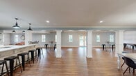 Located at 8003 SW Benaroya Ln in Huntsville, Alabama, this well-appointed single-floor space offers a generous, open layout designed for gathering and community use. Rich hardwood flooring flows seamlessly throughout, tying together each area with warmth and consistency. Crown molding accents the ceilings, lending a polished, refined character to every room.

The main living area features vaulted ceiling details, recessed lighting, and multiple windows that invite abundant natural light and offer views of the outdoor surroundings, including glimpses of a pool area. The open-plan layout connects the living space to the dining areas with ease, creating a welcoming, social atmosphere.

A standout feature is the kitchen bar area, anchored by a striking patterned tile backsplash, open floating shelves, white countertops, and a stone-clad bar base lined with black metal barstools. Industrial-style pendant lights hang overhead, adding a modern farmhouse aesthetic. A separate dining zone accommodates a large communal table beneath a cluster pendant light fixture, while a built-in counter workspace along a textured wallpapered wall provides additional seating. The open, connected design of the space makes it ideal for both casual gatherings and organized events.