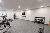 This fitness facility at 301 Sandwich St N in Amherstburg, Ontario, is a dedicated gym space designed to support a well-rounded workout routine. The room features a drop tile ceiling with recessed pot lighting that evenly illuminates the entire area, creating a bright and functional environment. The floor is finished with durable, patterned carpet tiles in a neutral grey tone, providing comfort underfoot while maintaining a clean, professional aesthetic. The walls are finished in a crisp white, contributing to the space's open and airy feel. The gym is comprehensively equipped with a variety of cardio machines, including a treadmill, elliptical trainer, stationary bike, and spin bike, catering to diverse cardiovascular training preferences. Strength training is well-represented with a multi-functional cable machine, a full rack of dumbbells, a flat weight bench, and additional free weight accessories. A wall-mounted flat-screen television with a dedicated shelf is centrally positioned for viewing during workouts. A vent-style window with vertical blinds allows for natural light, while a wall-mounted mini-split air conditioning unit ensures climate comfort year-round. The space is accessed through a dedicated entry door and is thoughtfully organized to maximize usability.