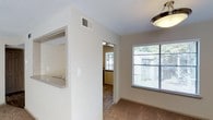 Located at 133 Lake Forest Drive in Daphne, Alabama, this single-story unit offers a clean, functional layout designed for comfortable everyday living. The interior features neutral-toned walls that create a bright, welcoming atmosphere throughout. The living area is finished with carpeted flooring and receives ample natural light through a large window fitted with horizontal blinds, offering views of the surrounding greenery. A flush-mount ceiling light fixture provides additional illumination. A pass-through window connects the living space to the kitchen area, offering a practical open-concept feel without fully removing the separation between rooms. The kitchen, visible through the pass-through and a connecting doorway, features countertops with a subtle speckled finish. Smooth, light-colored walls and white ceilings with a textured finish are consistent throughout the unit, contributing to a cohesive and tidy aesthetic. HVAC vents are integrated into the ceiling for efficient climate control. The single-floor design ensures easy accessibility, and the overall layout promotes a straightforward, low-maintenance living environment well-suited to the Daphne, Alabama community.
