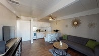 This single-floor space offers an open-concept design that integrates the kitchen and living area into one cohesive, functional layout. The kitchen is outfitted with white cabinetry, a gas range, a full-size refrigerator, and an array of countertop appliances, making it well-suited for everyday cooking. A peninsula with modern white bar stools serves as a natural divider between the kitchen and living space, providing both additional seating and a casual dining option. The living area features a contemporary gray sectional sofa accented with green throw pillows, centered around a round mosaic-top coffee table and a patterned area rug, creating a relaxed and inviting atmosphere. A ceiling fan with a wood-toned finish is mounted overhead, contributing to both airflow and the overall aesthetic. The space benefits from warm hardwood-style flooring that runs throughout, adding continuity and a sense of warmth. The walls are finished with a textured block-style treatment, while decorative sunburst accents lend a stylish, curated touch to the interior. The paneled white ceiling and open layout work together to create a bright, airy ambiance throughout the space.