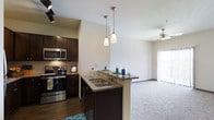 This single-floor residence offers a thoughtfully designed open-concept layout that seamlessly connects the kitchen and living area, creating a spacious and inviting atmosphere. The kitchen is a standout feature, showcasing rich dark cabinetry that contrasts beautifully with granite countertops and a full suite of stainless steel appliances, including a range, microwave, and refrigerator. A kitchen peninsula with a double basin sink serves as both a functional workspace and a casual dining or entertaining surface, illuminated by a pair of elegant pendant lights that add a warm, modern touch. Dark hardwood-style flooring anchors the kitchen area, while plush carpeting flows through the adjacent living space, creating a comfortable and welcoming transition between zones. The living area benefits from generous natural light streaming through large vertical-blind windows, and a ceiling fan enhances comfort year-round. Track lighting in the kitchen provides additional task illumination, ensuring the space is as practical as it is visually appealing. The clean, neutral wall palette throughout ties the design together, offering a versatile backdrop that complements the rich, contemporary finishes found across the space.