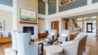 This thoughtfully designed community clubhouse makes a striking first impression with its soaring two-story lobby entrance, framed by floor-to-ceiling glass doors and expansive windows that flood the space with natural light. The main lounge area is anchored by a stunning stacked-stone fireplace with a wood mantel and a mounted flat-screen TV above, complemented by a curated arrangement of upholstered wingback chairs, sofas, and glass-topped coffee tables set atop area rugs over warm hardwood-style flooring. Deep teal accent walls and an elegant chandelier overhead add a refined, contemporary character to the double-height space. A separate lounge area offers floor-to-ceiling windows overlooking an outdoor terrace and surrounding community, creating a bright and inviting retreat. The kitchen features rich wood cabinetry, a light-blue tile backsplash, gray stone countertops, and stainless steel appliances, providing a fully functional space for gatherings. A private meeting or dining room includes a wood dining table with mid-century modern chairs, warm wall sconces, decorative framed artwork, and generous windows that bring in outdoor views. Throughout, the design balances comfort and sophistication with cohesive wood-toned flooring, neutral palettes, and carefully selected furnishings.