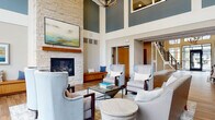 This thoughtfully designed community clubhouse makes a striking first impression with its soaring two-story lobby entrance, framed by floor-to-ceiling glass doors and expansive windows that flood the space with natural light. The main lounge area is anchored by a stunning stacked-stone fireplace with a wood mantel and a mounted flat-screen TV above, complemented by a curated arrangement of upholstered wingback chairs, sofas, and glass-topped coffee tables set atop area rugs over warm hardwood-style flooring. Deep teal accent walls and an elegant chandelier overhead add a refined, contemporary character to the double-height space. A separate lounge area offers floor-to-ceiling windows overlooking an outdoor terrace and surrounding community, creating a bright and inviting retreat. The kitchen features rich wood cabinetry, a light-blue tile backsplash, gray stone countertops, and stainless steel appliances, providing a fully functional space for gatherings. A private meeting or dining room includes a wood dining table with mid-century modern chairs, warm wall sconces, decorative framed artwork, and generous windows that bring in outdoor views. Throughout, the design balances comfort and sophistication with cohesive wood-toned flooring, neutral palettes, and carefully selected furnishings.