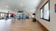 This generously sized fitness center offers a thoroughly equipped workout environment designed to support a wide range of exercise routines. The single-floor layout provides an open, unobstructed space with hardwood-style flooring throughout, creating a clean and functional atmosphere conducive to focused training.

The cardio zone features multiple stationary bikes, including Echelon and connected cycling bikes, elliptical machines, and treadmills, all positioned near large framed windows that invite natural light and views of the surrounding exterior. A wall-mounted flat-screen television adds an entertainment option for cardio sessions.

The strength training area is comprehensively outfitted with a multi-functional cable machine, a functional training rig with TRX suspension straps and battle ropes, a wall-mounted stall bar, a punching bag, a full dumbbell rack, adjustable benches, and a medicine ball collection. Large wall mirrors line portions of the space, enhancing the sense of openness while allowing for proper form checks during workouts.

Recessed lighting and ceiling fans ensure the space remains bright and comfortable throughout use. A dedicated entry vestibule with glass-paneled doors provides a clear transition into the facility. The warm, neutral wall tones and thoughtful layout make this fitness center an inviting and highly functional amenity.