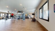 This generously sized fitness center offers a thoroughly equipped workout environment designed to support a wide range of exercise routines. The single-floor layout provides an open, unobstructed space with hardwood-style flooring throughout, creating a clean and functional atmosphere conducive to focused training.

The cardio zone features multiple stationary bikes, including Echelon and connected cycling bikes, elliptical machines, and treadmills, all positioned near large framed windows that invite natural light and views of the surrounding exterior. A wall-mounted flat-screen television adds an entertainment option for cardio sessions.

The strength training area is comprehensively outfitted with a multi-functional cable machine, a functional training rig with TRX suspension straps and battle ropes, a wall-mounted stall bar, a punching bag, a full dumbbell rack, adjustable benches, and a medicine ball collection. Large wall mirrors line portions of the space, enhancing the sense of openness while allowing for proper form checks during workouts.

Recessed lighting and ceiling fans ensure the space remains bright and comfortable throughout use. A dedicated entry vestibule with glass-paneled doors provides a clear transition into the facility. The warm, neutral wall tones and thoughtful layout make this fitness center an inviting and highly functional amenity.