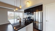 This single-floor residence offers a thoughtfully designed layout with a seamless blend of modern finishes and functional living spaces. The open-concept kitchen and living area serves as the heart of the home, featuring rich dark hardwood flooring that extends throughout the main living spaces. The kitchen is equipped with dark espresso cabinetry reaching toward the ceiling for ample storage, granite countertops in both dark and light tones, a subway tile backsplash, and a full suite of stainless steel appliances including a refrigerator, electric range, and built-in microwave. A central island with pendant lighting overhead provides additional prep space and casual seating. The spacious main living room is bathed in natural light through multiple windows fitted with blinds, creating a bright and welcoming atmosphere. The bathroom features a white vanity with an undermount sink, warm countertops, tile flooring, and warm vanity lighting above a large mirror. A generously sized walk-in closet with wire shelving offers abundant storage. An additional carpeted room with a stylish sliding barn door adds versatile living space. Throughout the residence, neutral wall tones and clean architectural lines contribute to a cohesive, contemporary aesthetic.
