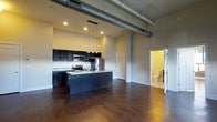 This single-floor unit showcases a distinctive industrial aesthetic defined by soaring ceilings with exposed metallic ductwork and structural columns, lending the space a loft-like character. The open-concept layout centers around a well-appointed kitchen featuring dark espresso cabinetry, stainless steel appliances including a refrigerator, range, and microwave, and a large central island with a light stone countertop that doubles as a prep and dining surface. Pendant lighting above the island adds warm, focused illumination to the cooking area, complementing the recessed lighting throughout. Rich, dark hardwood floors extend across the entire living area, grounding the space and contrasting cleanly with the neutral-toned walls. The generous open floor plan allows for flexible furniture arrangement in the living area. A bathroom is visible through an adjacent doorway, finished with standard fixtures. The combination of industrial overhead elements, modern finishes, and warm flooring creates a balanced, contemporary ambiance that is both functional and visually engaging.