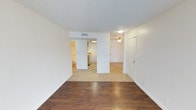 This single-story space showcases a practical and open layout, with distinct flooring materials delineating different functional zones. The main living area features rich, dark hardwood flooring that adds warmth and character, while the adjacent areas transition to light-toned tile, providing a clear visual separation between spaces. White walls throughout create a clean, bright atmosphere, complemented by a textured popcorn ceiling.

Structural half-walls and open passageways define the flow between areas without fully enclosing them, maintaining a sense of openness. A ceiling fan with a light fixture is visible in the adjoining room, providing both ventilation and ambient lighting. HVAC vents are integrated into the ceiling for climate control.

Glimpsed through the open passageways are a kitchen equipped with appliances and cabinetry, as well as a bathroom. A paneled white door on the right side of the space leads to an additional room or exterior access. The layout is straightforward and functional, designed to maximize usability across its connected spaces while maintaining a cohesive, neutral aesthetic throughout.