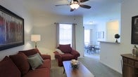 This single-floor residence offers a well-organized and comfortable living environment with a thoughtfully designed open layout. The living area is carpeted and anchored by a ceiling fan with integrated lighting, providing both comfort and airflow. Large windows dressed with blinds and curtains allow generous natural light to fill the space, creating a bright and welcoming atmosphere. The living room provides ample seating space and flows seamlessly into the dining area, which features a round table with chairs, making it ideal for everyday meals or casual gatherings. A partial wall with a pass-through counter connects the dining space to what appears to be a kitchen area, enhancing the open-concept feel while maintaining functional separation. The neutral wall tones throughout the space contribute to a clean, versatile aesthetic that suits a variety of furnishing styles. The consistent carpeting across the living and dining areas adds warmth and cohesion to the overall design. Flush-mount ceiling lighting in the dining area complements the ceiling fan fixture in the living room, ensuring the entire space is well-lit. Overall, this residence presents a practical and inviting single-floor layout suited for comfortable everyday living.