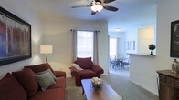 This single-floor residence offers a well-organized and comfortable living environment with a thoughtfully designed open layout. The living area is carpeted and anchored by a ceiling fan with integrated lighting, providing both comfort and airflow. Large windows dressed with blinds and curtains allow generous natural light to fill the space, creating a bright and welcoming atmosphere. The living room provides ample seating space and flows seamlessly into the dining area, which features a round table with chairs, making it ideal for everyday meals or casual gatherings. A partial wall with a pass-through counter connects the dining space to what appears to be a kitchen area, enhancing the open-concept feel while maintaining functional separation. The neutral wall tones throughout the space contribute to a clean, versatile aesthetic that suits a variety of furnishing styles. The consistent carpeting across the living and dining areas adds warmth and cohesion to the overall design. Flush-mount ceiling lighting in the dining area complements the ceiling fan fixture in the living room, ensuring the entire space is well-lit. Overall, this residence presents a practical and inviting single-floor layout suited for comfortable everyday living.