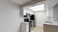 This single-floor space is highlighted by a well-appointed kitchen that combines modern functionality with a clean, contemporary aesthetic. The kitchen features crisp white shaker-style cabinetry offering ample storage, complemented by stainless steel appliances including a French door refrigerator, an electric range with a stainless steel over-the-range microwave. A standout feature of the kitchen is the recessed skylight panel in the ceiling, which floods the cooking area with bright, diffused light, creating an open and airy atmosphere. Light wood-look flooring runs throughout, adding warmth and continuity to the space. The countertops are finished in a clean white surface, providing generous workspace. The textured ceiling adds a subtle architectural detail consistent throughout the space. Neutral gray and white tones on the walls and surfaces contribute to a fresh, move-in-ready feel. The layout is efficient and thoughtfully arranged, with a countertop peninsula or bar area visible along one side, offering additional prep or dining space. Overall, the space presents a bright, modern environment well-suited for everyday living.