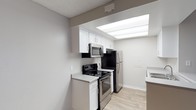 This single-floor space is highlighted by a well-appointed kitchen that combines modern functionality with a clean, contemporary aesthetic. The kitchen features crisp white shaker-style cabinetry offering ample storage, complemented by stainless steel appliances including a French door refrigerator, an electric range with a stainless steel over-the-range microwave. A standout feature of the kitchen is the recessed skylight panel in the ceiling, which floods the cooking area with bright, diffused light, creating an open and airy atmosphere. Light wood-look flooring runs throughout, adding warmth and continuity to the space. The countertops are finished in a clean white surface, providing generous workspace. The textured ceiling adds a subtle architectural detail consistent throughout the space. Neutral gray and white tones on the walls and surfaces contribute to a fresh, move-in-ready feel. The layout is efficient and thoughtfully arranged, with a countertop peninsula or bar area visible along one side, offering additional prep or dining space. Overall, the space presents a bright, modern environment well-suited for everyday living.