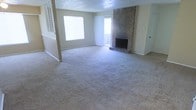 This single-floor space centers around a generously sized living room that offers an open, airy feel. The room is finished with wall-to-wall carpet in a neutral tone, providing a comfortable and cohesive foundation throughout. A standout feature is the full-height brick fireplace, which serves as a bold focal point along one wall, adding a classic and cozy architectural element to the space.

Natural light flows into the room through a large window on one side, while a set of sliding glass doors with sheer curtains opens to an outdoor area, enhancing the sense of openness and connectivity to the exterior. The combination of these light sources keeps the interior bright and welcoming.

The layout includes a partial dividing wall or column near the entryway, subtly defining zones within the open floor plan without sacrificing the overall sense of spaciousness. The walls are finished in a soft, neutral tone, and the textured ceiling adds a layer of traditional character to the space. Overall, the living area presents a functional and classic design well-suited to comfortable everyday living.
