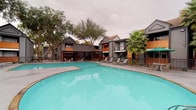 This residential community showcases a thoughtfully designed outdoor environment that blends classic and contemporary architectural styles. The grounds feature two distinct pool areas: one set against a backdrop of dark-painted wood-sided buildings with warm timber accents, and another framed by traditional multi-story brick structures with steeply pitched rooflines and bay windows. Both pools are generously sized, with clear turquoise water and surrounding pool decks furnished with lounge chairs for relaxation.

The brick buildings display rich red-and-tan masonry detailing, large grid-paned windows, and private balconies with wrought iron railings, giving the community a refined, upscale character. Mature trees — including live oaks and tall palm trees — are thoughtfully integrated throughout the grounds, providing natural shade and softening the hardscape.

A spacious paved courtyard connects the pool areas, featuring outdoor seating arrangements with umbrella-shaded tables and chairs, ideal for social gatherings. Well-maintained landscaping with manicured shrubs, ground cover, and ornamental plantings lines the building perimeters, enhancing the community's curb appeal. A clubhouse or amenity building with floor-to-ceiling windows provides an indoor-outdoor connection to the pool, completing this inviting and cohesive community environment.