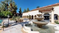 This single-story property showcases a distinctive Mediterranean Revival architectural style, characterized by warm stucco exteriors, terracotta tile roofing, and a series of elegant arched colonnades. The covered walkways feature repeating arched openings supported by substantial stucco columns, creating shaded passages that connect various areas of the property while maintaining an open, airy feel.

At the heart of the outdoor space stands a prominent multi-tiered stone fountain, serving as a central focal point within a sun-drenched courtyard finished with neutral stone pavers. The courtyard offers outdoor seating areas framed by mature olive trees and desert-adapted landscaping, providing a comfortable gathering space beneath the clear blue sky.

The resort-style swimming pool area is generously proportioned and lined with lounge chairs, set against a backdrop of towering date palms and dense, mature trees that provide natural shade and privacy. Landscaped planters and decorative lamp posts punctuate the pool deck, contributing to the property's polished outdoor ambiance.

Interior-facing arched windows with decorative transom detailing allow natural light into the corridors, while the textured stucco wall finishes reinforce the cohesive Mediterranean aesthetic throughout both the interior and exterior spaces.