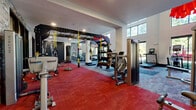 This expansive fitness room in Birmingham, Alabama, offers a comprehensive workout environment designed to accommodate a wide range of exercise needs. The space is architecturally striking, featuring soaring double-height ceilings and large floor-to-ceiling windows that flood the interior with natural light while providing views of surrounding greenery. A mezzanine-level railing overlooks the main floor, adding a sense of vertical scale and openness to the overall design.

The flooring throughout consists of modular carpet tiles in a bold pattern of red, gray, and charcoal, creating distinct functional zones across the space. Stone accent walls add texture and warmth, complementing the contemporary industrial aesthetic defined by exposed black steel framing and recessed lighting.

Equipment is extensive and well-organized. A dedicated cardio zone features Matrix treadmills and elliptical machines positioned along the windowed wall. A functional training area includes a multi-station suspension rig with TRX straps and a heavy bag. Strength training is supported by a selection of Matrix selectorized machines, cable systems, adjustable benches, and a rack stocked with medicine balls and kettlebells. A mirrored section provides space for stretching or individual training. The overall ambiance is energetic and modern, well-suited for a full-service fitness experience.
