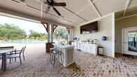 Located at 100 Pinckney Marsh Lane in Port Royal, South Carolina, this single-story community amenity pavilion offers an expansive covered outdoor gathering space designed for recreation and relaxation. The structure is defined by its striking white shiplap exterior, warm wood-accented timber brackets, and a coffered ceiling with multiple ceiling fans that ensure comfortable airflow throughout. Rustic brick pavers span the entire floor, lending a classic Lowcountry charm to the space.

The pavilion is thoughtfully organized into several distinct zones. A generously sized lounge area features wicker sectional sofas and armchairs accented with blue throw pillows, arranged around a central coffee table. Adjacent to the lounge, a well-equipped outdoor kitchen includes white cabinetry, a central island with bar seating, a wall-mounted flat-screen television, and beverage refrigerators. Dining tables with chairs provide additional seating for larger gatherings. Entertainment options include a shuffleboard table, ping-pong table, and a beverage vending machine.

Large glass French doors connect the pavilion to an enclosed interior clubhouse, while open-air columns frame sweeping views of the resort-style swimming pool and lush, tree-lined grounds beyond.