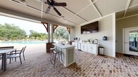 Located at 100 Pinckney Marsh Lane in Port Royal, South Carolina, this single-story community amenity pavilion offers an expansive covered outdoor gathering space designed for recreation and relaxation. The structure is defined by its striking white shiplap exterior, warm wood-accented timber brackets, and a coffered ceiling with multiple ceiling fans that ensure comfortable airflow throughout. Rustic brick pavers span the entire floor, lending a classic Lowcountry charm to the space.

The pavilion is thoughtfully organized into several distinct zones. A generously sized lounge area features wicker sectional sofas and armchairs accented with blue throw pillows, arranged around a central coffee table. Adjacent to the lounge, a well-equipped outdoor kitchen includes white cabinetry, a central island with bar seating, a wall-mounted flat-screen television, and beverage refrigerators. Dining tables with chairs provide additional seating for larger gatherings. Entertainment options include a shuffleboard table, ping-pong table, and a beverage vending machine.

Large glass French doors connect the pavilion to an enclosed interior clubhouse, while open-air columns frame sweeping views of the resort-style swimming pool and lush, tree-lined grounds beyond.