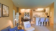 This single-floor residence welcomes you with a seamlessly connected open-concept layout that integrates the living, dining, and kitchen areas into one cohesive, comfortable space. The interior is finished in warm neutral tones — soft beige walls and light carpet flooring — that create an inviting and airy ambiance throughout.

The living area is thoughtfully arranged, featuring cozy seating with blue and white coastal accents, including wicker furniture and plaid throw pillows that reinforce a relaxed, beach-inspired aesthetic. A decorative arrangement of white florals adds an elegant touch to the corner of the room.

The dining area sits centrally within the open plan, anchored by a round dark-wood table with wicker chairs. A white display cabinet nearby showcases coastal decorative pieces such as starfish, lending the space a curated, seaside charm. A paddle mounted on the wall further enhances the nautical theme.

The kitchen is well-appointed with white cabinetry, white appliances, and a breakfast bar with seating, making it both functional and social. Track lighting and pendant fixtures provide warm, layered illumination throughout the combined living and dining areas. The overall design balances practicality with a cohesive coastal aesthetic, making the space feel comfortable and welcoming.