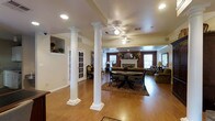 This single-story space showcases a thoughtfully designed open floor plan that seamlessly connects the dining and living areas. Prominent white decorative columns with detailed bases and capitals serve as elegant architectural dividers, adding a classic, formal character to the interior without fully separating the spaces.

The flooring throughout is warm-toned hardwood, contributing to a cohesive and inviting atmosphere. Ceiling fans with integrated lighting are positioned across the ceiling, providing both comfort and illumination, complemented by recessed lighting throughout.

The dining area features a dark wood table set atop a patterned area rug, anchoring the space with a traditional aesthetic. Crown molding runs along the ceiling perimeter, adding a refined finishing detail.

The living area at the rear is defined by rich, deep red accent walls and centers around a classic white fireplace with a mantel, above which a flat-screen television is mounted. Multiple windows flank the fireplace, allowing natural light to fill the room. A separate room accessible through French doors is visible, adding additional functional space to the layout.