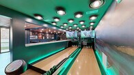 Located at 4521 Landmark Dr in Raleigh, NC, this single-floor entertainment venue is a boldly designed space that combines recreation and social amenities in one cohesive environment. The centerpiece is a private bowling alley featuring two full lanes illuminated by striking green LED lighting along the lane gutters, a vivid green ceiling with recessed can lights, and large projection screens displaying dynamic visuals at the pin end — creating an immersive, arcade-like atmosphere. Adjacent to the lanes is a dedicated bar and lounge area finished in deep teal walls and a reflective ceiling tile system, with a long white-topped bar counter lined with sleek black barstools. Colorful abstract artwork adorns the walls, and herringbone-patterned light wood flooring ties the space together with a warm yet modern aesthetic. A separate lounge area offers an intimate retreat with rich teal walls, a glossy teal ceiling, herringbone flooring, and curated furnishings including a tufted sofa, vibrant yellow accent chairs, a geometric coffee table, and layered area rugs. Wall sconces and framed artwork complete the sophisticated ambiance. The overall design merges entertainment, hospitality, and interior artistry into a one-of-a-kind private venue experience.