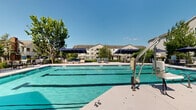 Located at Fonthill Court in Langhorne, PA, this outdoor pool and amenity area serves a three-story residential community. The centerpiece is a generously sized swimming pool with clearly marked lap lanes, making it suitable for both recreational swimming and structured exercise. The pool's vivid turquoise water and clean, light-colored deck create a bright, inviting atmosphere on sunny days.

The surrounding deck is well-appointed with lounge chairs, tables, and seating areas positioned throughout, offering ample space for sunbathing and socializing. Multiple navy blue shade canopies are strategically placed around the pool perimeter, providing relief from the sun while maintaining an open, airy feel. An accessibility lift is present at the pool's edge, ensuring ease of access for all users.

Adjacent to the pool, a charming pavilion with a pitched roof provides additional covered seating and gathering space. A dedicated clubhouse building sits at one end of the pool area, featuring large arched windows and glass double doors accessed via a short staircase with white railings and high-visibility step edges. Lush mature trees frame the space, adding greenery and natural shade. The three-story residential buildings visible in the background complete the well-maintained, community-focused setting.