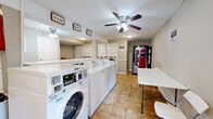 This communal laundry facility is a compact, single-floor space thoughtfully designed to accommodate the laundry needs of multiple users. The main area is lined with a row of full-sized, commercial-grade top-loading and front-loading washing machines alongside stacked dryer units, providing ample capacity for simultaneous use. Ceiling fans are mounted overhead, promoting air circulation throughout the space, which is finished with durable, easy-to-clean beige tile flooring throughout.

Decorative "Wash," "Dry," and "Fold" signage is displayed on the walls, guiding users through the laundry process in a clear and organized manner. A dedicated folding station features white cabinetry with a stone-patterned countertop surface, offering a practical workspace for sorting and folding laundry. A white table with seating provides additional space for residents to wait or attend to their laundry.

A Pepsi vending machine is conveniently positioned near the entrance area, adding a welcome amenity for users. The facility is equipped with a fire alarm safety system and clearly posted instructional signage. Flush-mounted and ceiling fan lighting fixtures ensure the space is well-lit throughout. The neutral, light gray walls and white fixtures create a clean, bright atmosphere that is both functional and welcoming.