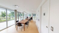 Located at 4277 Norland Ave in Burnaby, BC, this single-floor communal space is thoughtfully designed to serve as a shared kitchen and dining area. The layout is clean and functional, with a bright, open atmosphere achieved through an all-white color palette, recessed lighting, and light-toned hardwood flooring throughout.

The kitchen runs along one wall in a galley-style configuration, outfitted with ample upper and lower cabinetry in a two-tone white and natural wood finish. It features a double stainless steel sink, an electric range with four coil burners, a dishwasher, a full-size refrigerator, and generous counter space — making it well-suited for group meal preparation.

The adjoining dining area is the heart of the space, anchored by wooden dining tables and mixed seating. Exposed concrete columns add an architectural character to the room, complementing the modern design. The standout feature is the expansive floor-to-ceiling glazing along the exterior wall, allowing an abundance of natural light to flood the interior while offering open views of the surrounding landscaped outdoor area and streetscape. A sliding glass door provides direct access to the exterior patio. The overall ambiance is welcoming, airy, and well-suited for communal dining and social interaction.