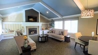 This single-floor space centers around a stylish living area distinguished by its striking vaulted ceiling, finished in a bold blue-gray tone that adds depth and visual interest. White trim lines the ceiling's angles, creating a clean architectural contrast. A focal fireplace with a tiled surround anchors one wall, topped by a wall-mounted flat-screen TV set within a defined architectural niche. Large windows along the opposite wall allow generous amounts of natural light to flood the interior, enhancing the open, airy feel of the space. The living area is furnished with a pair of neutral-toned sofas arranged around a dark, round coffee table, complemented by coordinating side tables and decorative accents including floral arrangements and textured throw pillows. A patterned area rug grounds the seating arrangement and adds warmth underfoot. Built-in window seating provides additional functional space near the windows, offering a cozy nook for relaxing. Lush indoor plants contribute a fresh, natural element to the overall ambiance. The design balances a warm, welcoming character with refined architectural details throughout.