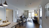 This single-floor residence offers an open-concept layout that seamlessly connects the kitchen, dining, and living areas into one cohesive space. The kitchen features granite countertops, providing a clean transition into the dining area, which accommodates a round glass-top table with dark wooden chairs. Rich hardwood flooring runs throughout, adding warmth and continuity across the main living spaces.

The living area is well-proportioned, offering room for a full sofa arrangement centered around a glass coffee table and a patterned area rug. A floor lamp provides ambient lighting, complementing the abundant natural light that flows in through large windows dressed with neutral curtains. These windows frame views of the surrounding exterior and lead to a balcony, enhancing the sense of openness.

A sitting nook near the windows provides an additional relaxation spot, complete with an accent chair. White walls and standard-height ceilings keep the space feeling bright and airy throughout. A doorway visible from the main area suggests access to a bedroom, indicating a functional and well-organized floor plan overall.