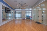 This single-level fitness facility offers a thoughtfully designed layout with multiple dedicated workout zones. The centerpiece is a spacious studio room with warm hardwood flooring, wall-to-wall framed mirrors, and a mounted flat-screen television for guided workouts. A horizontal ballet barre runs the length of the mirrored wall, making the space well-suited for barre, yoga, dance, or group fitness classes. The studio's tray ceiling with recessed lighting adds architectural interest and a sense of height, while neutral gray walls and white trim create a clean, polished aesthetic.

Adjacent to the studio is a glass-enclosed racquetball court, enclosed by full-height tempered glass panels and chrome hardware that allow clear sightlines from the surrounding hardwood-floored common area. Natural light filters in through large windows along the perimeter, brightening the space considerably.

A dedicated equipment area houses a rack of stability and medicine balls alongside a full dumbbell rack, positioned near floor-to-ceiling windows that allow ample natural light and views of the surrounding exterior. Throughout the facility, the consistent use of hardwood flooring, neutral tones, and abundant lighting creates a cohesive, professional, and inviting environment suited to a wide range of fitness activities.