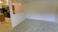 This single-floor space features a spacious living area finished with wall-to-wall carpeting in a neutral tone, providing a soft, understated foundation throughout. A standout architectural detail is the decorative half-wall partition fitted with white-painted wooden balusters, which visually separates the living area from the adjacent kitchen while maintaining an open, connected feel. The kitchen is partially visible beyond the divider and appears to include cabinetry and countertop space. The living area walls are painted in a light, neutral cream tone, contributing to a clean and bright atmosphere. Multiple electrical outlets are positioned along the walls, offering practical placement for furniture and appliances. The overall layout is straightforward and open, making the space adaptable for a variety of furnishing arrangements. The combination of the soft carpeting, light wall tones, and the distinctive baluster partition gives the space a classic, understated residential character suited for comfortable everyday living.