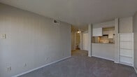 This single-story unit is organized into a practical, efficient layout as seen in the floor plan. The living area is generously proportioned, featuring neutral gray carpet, smooth gray walls, a textured ceiling, and a ceiling-mounted HVAC vent for climate control. A sliding glass door with vertical blinds allows natural light to fill the space and provides access to an outdoor area. The entry door is positioned adjacent to the sliding door, creating a welcoming entryway.

The galley-style kitchen is accessible via a pass-through peninsula and features white cabinetry offering ample storage, granite-style countertops, a stainless steel sink, an electric range with oven, a full-size refrigerator, a dishwasher, and wood-look vinyl flooring — a practical contrast to the carpeted living areas.

The bedroom is a comfortable, carpeted space with neutral walls, a ceiling fan with light fixture, and a window providing natural light and exterior views. The textured ceiling is consistent throughout the unit.

The bathroom is efficiently designed, featuring a tub-shower combination with tiled surround, a toilet, a white vanity with granite-style countertop and integrated sink, a large mirror, and wood-look vinyl flooring matching the kitchen.