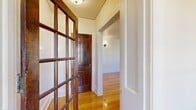 This single-floor home offers a well-organized layout encompassing three bedrooms, one bathroom, two living areas, a dining room, kitchen, entrance, and two hallways. Warm-toned hardwood floors run consistently throughout the main living and bedroom areas, contributing to a cohesive and inviting aesthetic across the home.

The entry is framed by a distinctive multi-pane French door with rich wood detailing, setting a classic architectural tone that continues throughout. The hallways connect rooms efficiently, with traditional wood-panel interior doors and flush ceiling light fixtures providing functional elegance.

One of the living rooms serves as a central gathering space, highlighted by a wood-burning fireplace with a white surround, black tile hearth, and flanking built-in shelving units — a notable focal point of the home. Multiple grid-pane windows across the living and bedroom areas allow generous natural light and views of outdoor greenery.

The bedrooms feature the same polished hardwood flooring and bright white walls, with windows providing natural light and outdoor views. The kitchen includes warm wood cabinetry, light countertops, tile flooring, and a back door providing additional access. The bathroom features a full tub-shower combination with white tile surround and mosaic tile flooring.