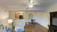 This single-floor space features an open-concept design that seamlessly connects the living, dining, and kitchen areas into one cohesive environment. The living area is anchored by a ceiling fan with an integrated light fixture, providing both airflow and ambient lighting throughout the space. Hardwood-style flooring runs continuously across the entire layout, lending warmth and visual consistency from room to room.

The kitchen is positioned at the rear of the open floor plan, separated by a breakfast bar counter that doubles as a transitional element between cooking and living spaces. Honey-toned wood cabinetry lines the kitchen walls, complemented by a black refrigerator. A decorative "Cucina" sign adds a personalized touch above the cabinetry.

The dining area sits adjacent to the kitchen and living space, defined by a patterned area rug beneath a dining table and upholstered chairs. A striking botanical wall decal serves as a focal point on the accent wall, adding an artistic element to the room.

The living area features patterned upholstered seating and flows naturally into the dining space, creating a comfortable and functional environment suitable for everyday living and entertaining.