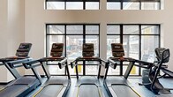 This contemporary fitness center is designed to deliver a premium workout experience within a bright, open, and thoughtfully organized single-floor space. Soaring ceilings and expansive floor-to-ceiling windows with black steel frames flood the interior with natural light and provide views of an outdoor amenity area, including a pool and courtyard spaces visible from the cardio zone.

The facility is accessed through sleek glass double doors framed in black steel, opening into a spacious, airy interior. The main workout floor is defined by polished structural columns, large wall-mounted mirrors, and smooth, durable flooring throughout. Cardio equipment is prominently featured along the window-facing walls, with multiple treadmills and elliptical machines arranged to take full advantage of the outdoor views. A dedicated strength training area houses a robust selection of free weights, a full dumbbell rack, adjustable benches, and a multi-functional cable machine system. A separate open studio area with mirrored walls offers ample space for stretching, group exercise, or functional training. Wall-mounted flat-screen televisions and recessed linear lighting contribute to a clean, modern aesthetic. The overall ambiance is energetic yet refined, combining professional-grade equipment with contemporary architectural design.
