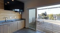 This contemporary single-floor space is thoughtfully designed with clean lines and modern finishes throughout. The interior showcases a well-appointed kitchenette area featuring warm wood-grain cabinetry paired with a dark countertop, complemented by a natural wood tile backsplash that adds texture and visual interest to the space. A wall-mounted flat-screen TV is positioned above the cabinetry, making the area both functional and entertainment-ready. The flooring consists of sleek wood-look planks that run throughout, contributing to the cohesive, modern aesthetic. A large sliding glass door serves as a prominent architectural feature, flooding the interior with natural light and providing a seamless transition to the outdoor deck. The deck is generously sized, finished with warm wood decking, and furnished with a dining table and chairs, making it ideal for outdoor relaxation or entertaining. A cable-rail perimeter allows for unobstructed views of the surrounding neighborhood and lush tree canopy beyond. String lights are visible overhead on the deck, adding an inviting ambiance to the outdoor area. The overall design balances modern minimalism with warm natural materials, creating a functional and welcoming environment.